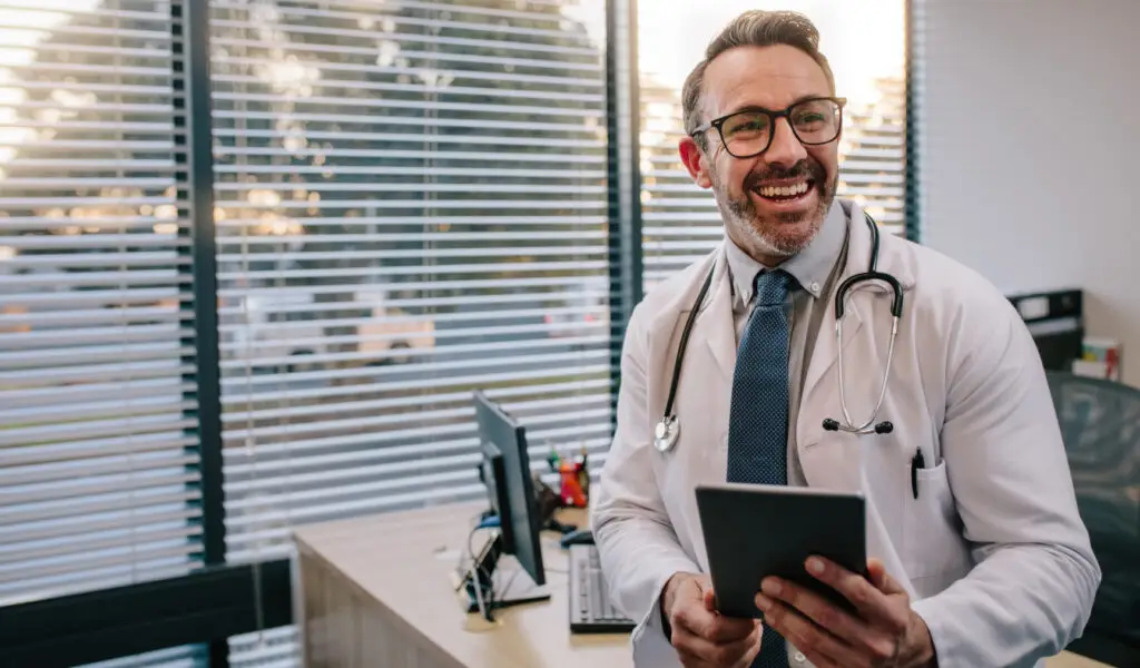 Smiling doctor using a tablet in a modern office, showcasing the role of digital healthcare, e-consults, and curbside consultations in patient care.